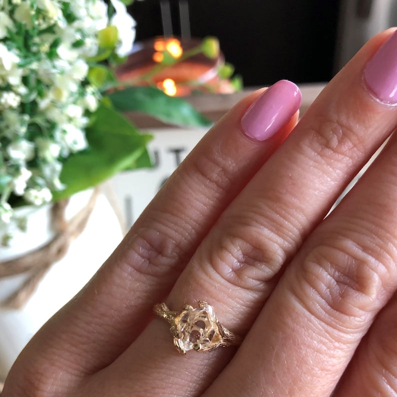 Hand wearing a yellow gold ring with pink nail polish in front of flowers