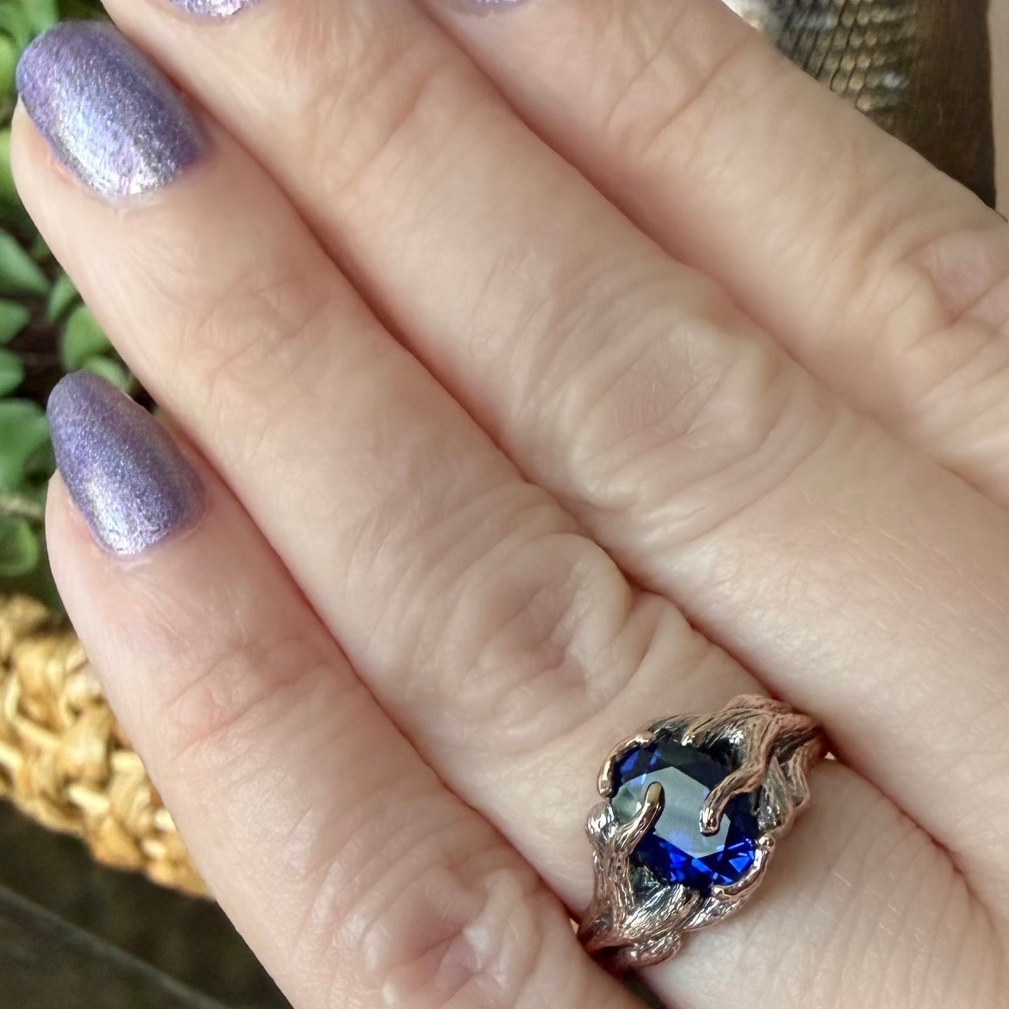 Hand with glittery purple nails wearing a ring with a blue gemstone.