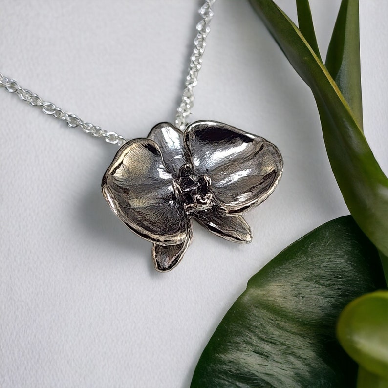 Silver orchid pendant necklace on a white background with green leaves.