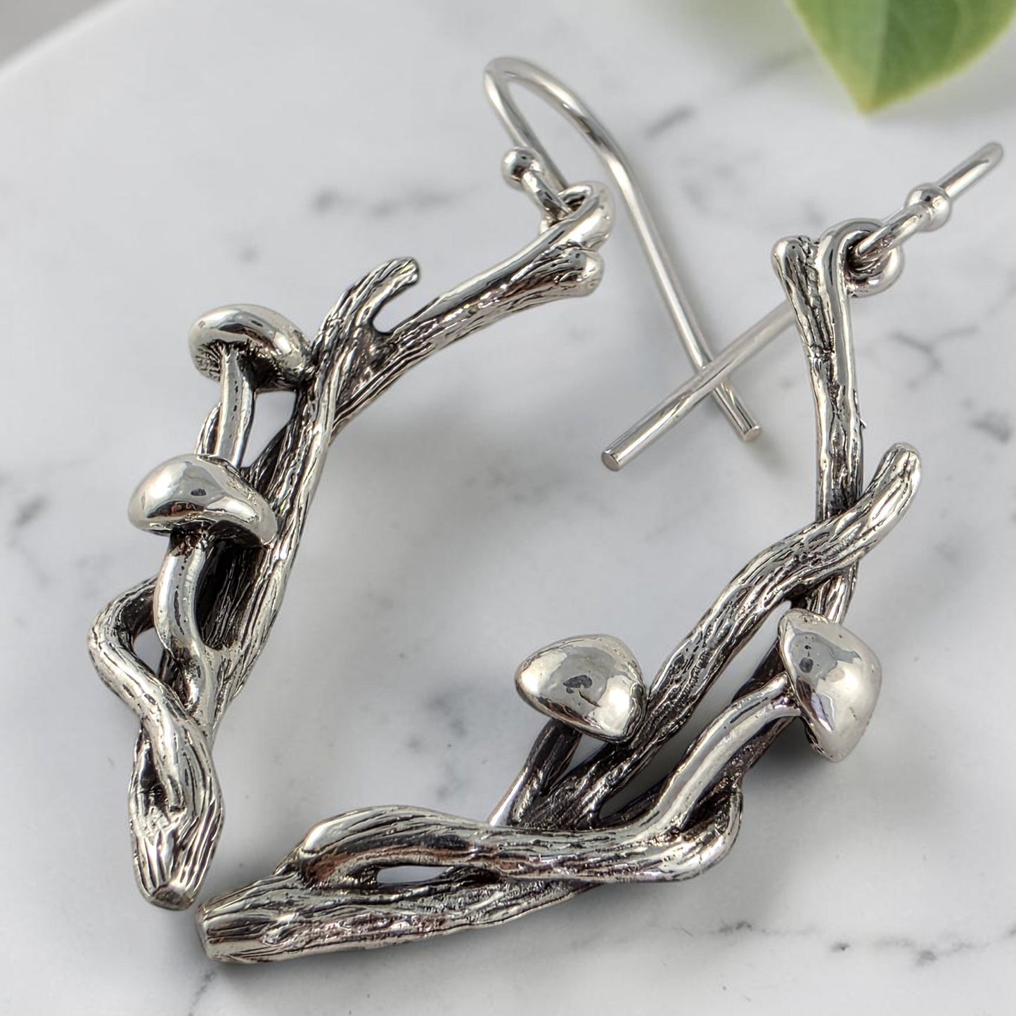 Silver branch-shaped earrings with mushrooms on a white surface with green leaves in the background