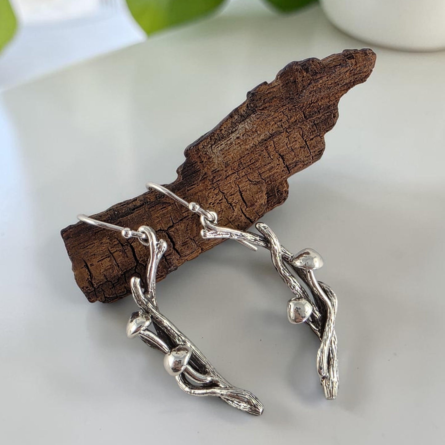 Silver branch-shaped earrings with mushrooms on a white surface with green leaves in the background