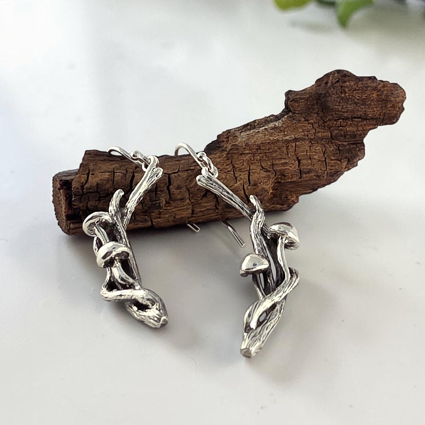 Silver chain earrings on a white surface with a piece of wood and greenery in the background