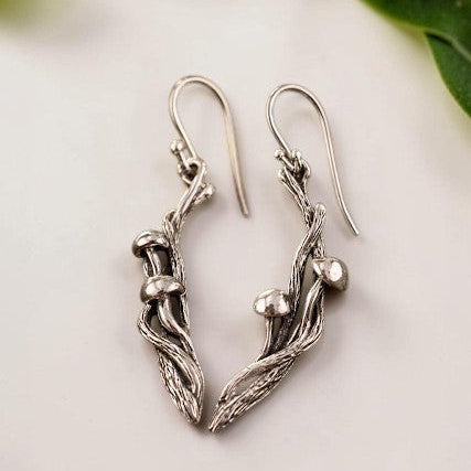 Silver branch-shaped earrings with mushrooms on a white surface with green leaves in the background