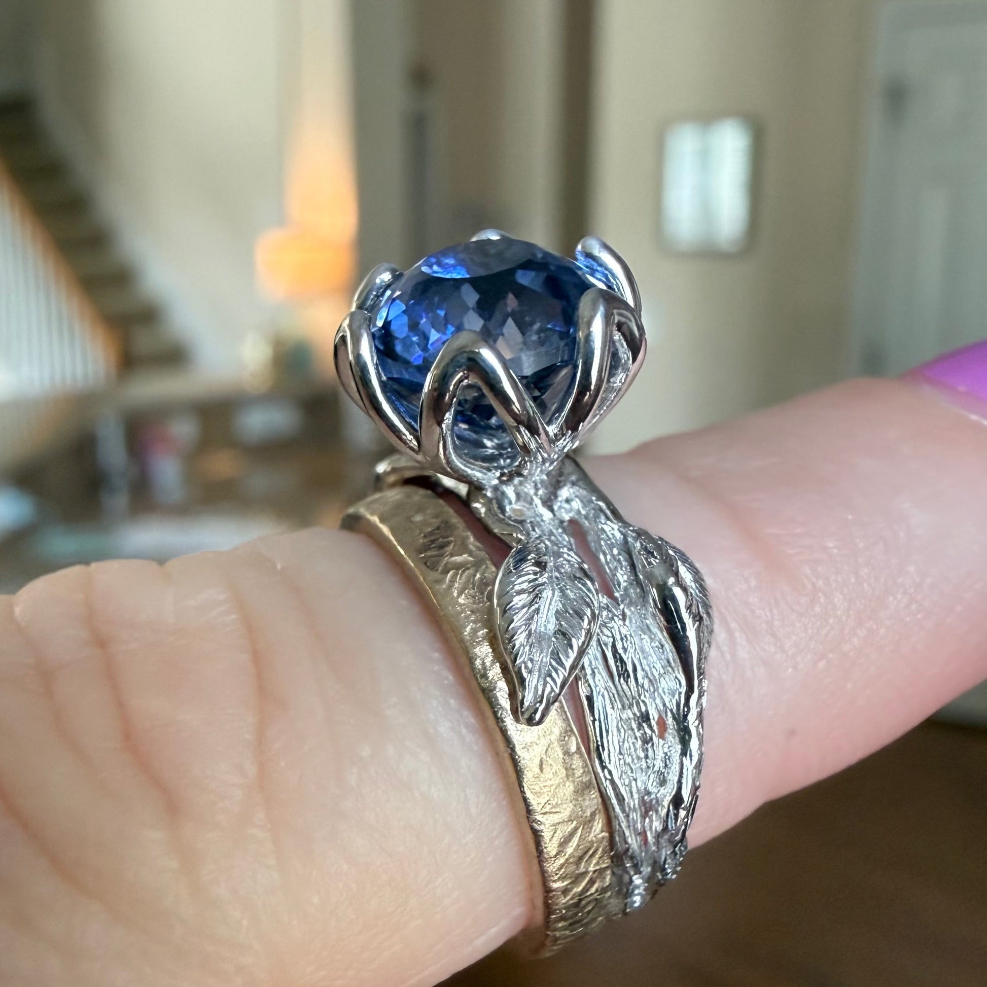 A close-up of a hand wearing a silver ring with a large faceted blue gemstone and textured leaf-like bands, set against a blurred indoor background.